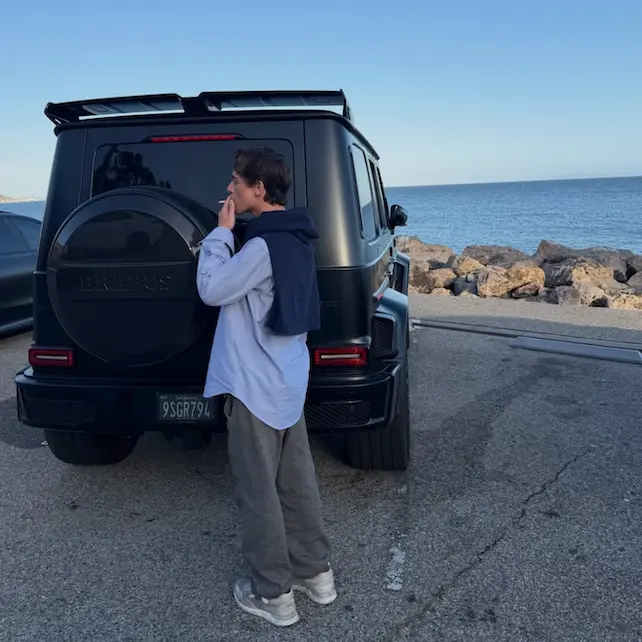 Hudson Cosper smokes a cigarette behind his G-Wagon with the ocean in the background.